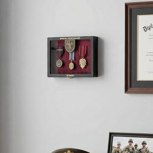 May include: A display of military memorabilia including medals in a shadow box, a framed certificate, a framed photograph of soldiers, and a military hat on a wooden desk. The shadow box has a red velvet backing.