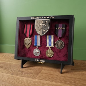 May include: A black display case containing military medals and insignia. The case has a red velvet interior and is supported by two black legs. The medals have various coloured ribbons and metal designs. The text "ВОЛЕЮ ТА ЗАЛІЗОМ" is at the top.