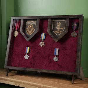 May include: A dark wooden display case with a burgundy velvet backing, displaying medals and crests. The case has a framed design with angled supports. The medals have various coloured ribbons.