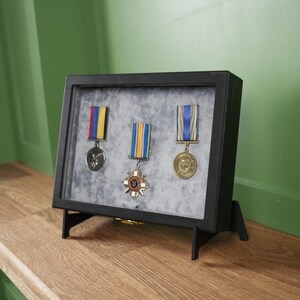 May include: A black framed display case holding three medals with colorful ribbons. The medals are set against a gray background. The case is displayed on a wooden surface against a green wall.