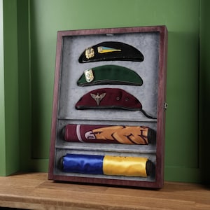 May include: A wooden display cabinet with a glass front, containing military berets and rolled banners. The berets are black, green, and maroon, each with a badge. The banners are rolled, displaying various colours and designs.