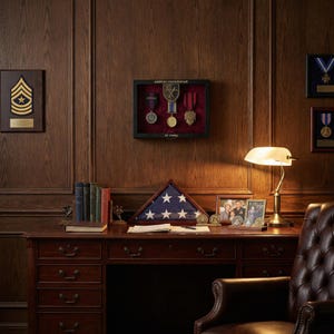 May include: A mahogany desk with a leather armchair and a lamp. The desk displays a folded American flag, framed photographs, and military medals. A display case with medals hangs on the wood-panelled wall, along with a military insignia.