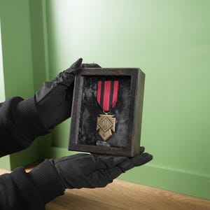 May include: A framed military medal with a red and black striped ribbon. The medal is gold-colored and has a detailed design. The frame is dark wood, and the background is a muted green.