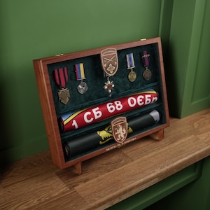 Op de afbeelding: Een houten vitrine met een donkergroen fluwelen interieur, met militaire medailles en een opgerolde banner. De vitrine bevat medailles in rood, blauw en goud, en een banner met Cyrillische tekst.