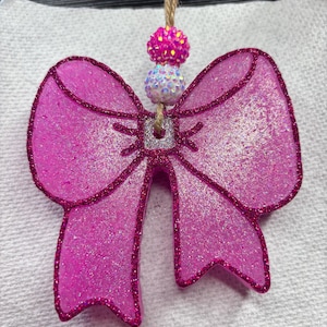 May include: A decorative pink glitter bow-shaped ornament with a sparkly outline and a jute string. The bow features a silver centre and two decorative beaded spheres in pink and white. This festive decoration is perfect for the holidays.