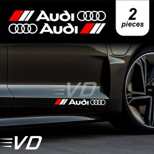 Peut inclure: Un ensemble de deux autocollants blancs Audi avec des accents rouges et noirs, présentant le logo et le texte Audi. Le design inclut un élément rayé rouge et noir stylisé. Les autocollants sont appliqués sur le panneau latéral d'un véhicule gris.