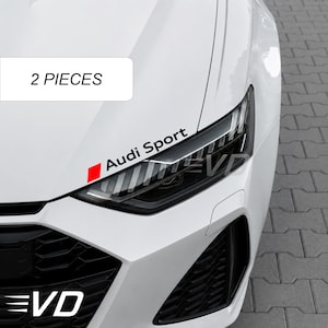 Può includere: Un cofano e un faro di un'auto bianca con un adesivo nero Audi Sport con un quadrato rosso. L'adesivo è posizionato sopra il faro. Un'etichetta rettangolare bianca con il testo nero 2 PIECES è visibile nell'angolo in alto a sinistra.