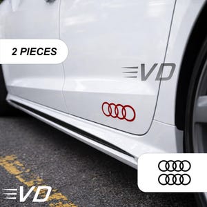 Op de afbeelding: Een set van twee rode vinylstickers met het vierringige Audi-logo aangebracht op de zijkant van een witte auto. De tekst 2 PIECES is zichtbaar in een wit kader en een aparte afbeelding toont twee zwarte ringlogo's verticaal op elkaar.