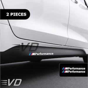 Op de afbeelding: Een set van twee MPerformance vinylstickers voor de zijskirts van een auto. De stickers bevatten de witte tekst MPerformance voorafgegaan door een logo met schuine blauwe, paarse en rode strepen, aangebracht op de zwarte dorpel van een witte auto.