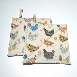 May include: Two square quilted pot holders with a cream background. The pot holders are decorated with colorful chicken patterns. Each chicken has a unique design. The pot holders have a small loop for hanging.