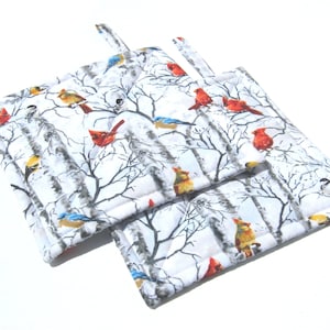 May include: A pair of white quilted pot holders with a wintery forest scene featuring red cardinals, blue jays, and yellow birds on branches.