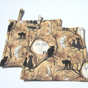 May include: Two beige quilted pot holders with a black cat and moon design. The pot holders have a loop for hanging.