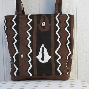 May include: A brown tote bag with a repeating pattern of vertical stripes and white zigzag lines. The bag features a dark brown base and handles, with small shell accents. The design has a tribal or African-inspired aesthetic.