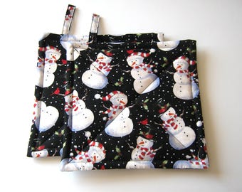 May include: Two black quilted pot holders with a snowman pattern. The snowmen are wearing red and white hats and scarves. The background is black with white polka dots and green holly leaves.