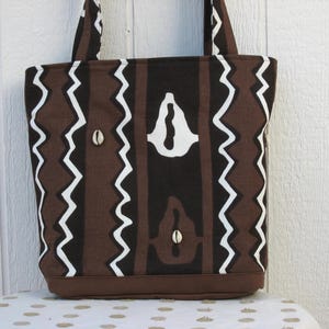 May include: A brown and white tote bag with a geometric pattern. The bag features a repeating design of brown vertical stripes, white zigzags, and white shapes. The bag has brown handles and a brown base, and includes two small shell accents.