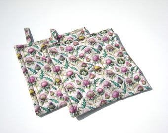 May include: Two square pot holders with a floral pattern featuring pink thistles, green leaves, and yellow accents on a white background. Each pot holder has a small fabric loop for hanging.