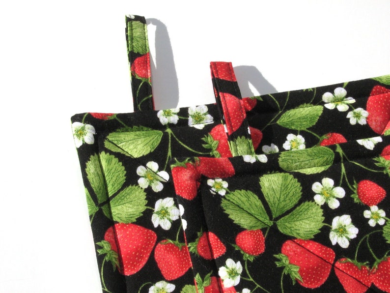 May include: Black and white pot holder with a strawberry print. The pot holder has two loops for hanging and is made of fabric.