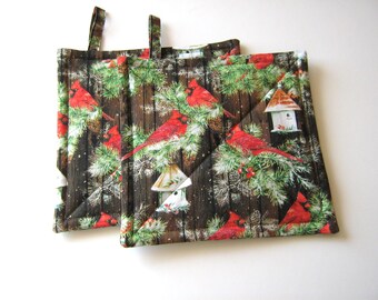 May include: Two quilted pot holders with a winter woodland theme. The pot holders feature a brown wood background with red cardinals, pine branches, and white birdhouses.