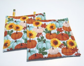 May include: Two quilted pot holders with a blue and white gingham background. The pot holders feature a pattern of pumpkins, sunflowers, and fall leaves.