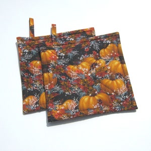 May include: Two brown and black pot holders with a fall-themed print of orange pumpkins and white flowers on a dark background.