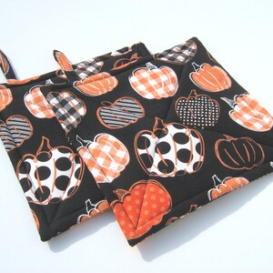 May include: Two black quilted pot holders with a pumpkin pattern in orange and white. The pumpkins have different patterns, including polka dots, gingham, and stripes.