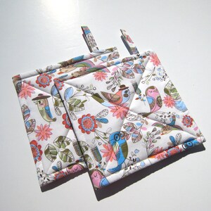 May include: Two white quilted pot holders with a colorful bird and floral pattern. The pot holders have a loop for hanging.