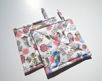 May include: Two white quilted pot holders with a colorful bird and floral pattern. The pot holders have a loop for hanging.