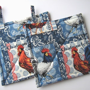 May include: Two square quilted pot holders with a blue and white rooster print. The fabric features roosters, paisley patterns, and the words "Bon Matin" and "Chateau". Each pot holder has a fabric loop for hanging.