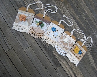 May include: Five rectangular brown cardstock tags featuring white lace trim, crochet accents, and fabric flowers. Each tag includes a white string loop at the top for hanging. The embellishments include small pearls, green leaves, and various colored floral shapes.