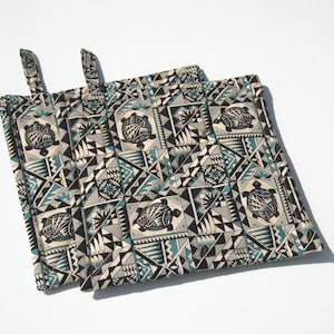 May include: Two square pot holders with hanging loops. The fabric features a repeating pattern of turtles, triangles, and geometric shapes in black, teal, and gray on a cream background. The pot holders are designed for kitchen use.