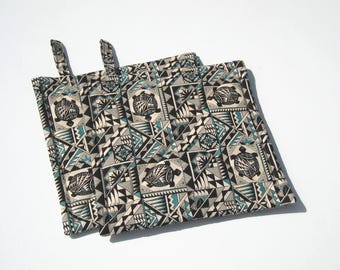 May include: Two square pot holders with hanging loops. The fabric features a repeating pattern of turtles, triangles, and geometric shapes in black, teal, and gray on a cream background. The pot holders are designed for kitchen use.