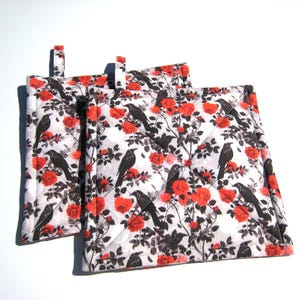 May include: Two square quilted pot holders with a white background, featuring a pattern of black crows and red roses. Each pot holder has a small fabric loop for hanging.