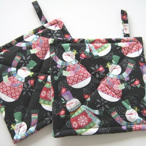 May include: Two black quilted pot holders with a snowman pattern. The snowmen are wearing colorful scarves and hats. The pot holders have a loop for hanging.
