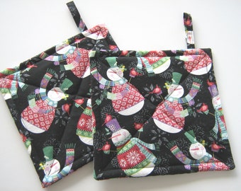 May include: Two black quilted pot holders with a snowman pattern. The snowmen are wearing colorful scarves and hats. The pot holders have a loop for hanging.