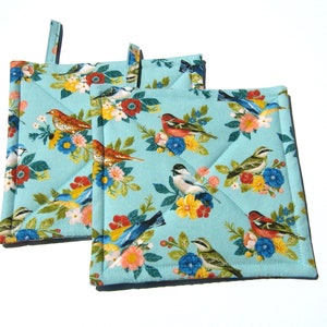 May include: Two blue quilted pot holders with a floral pattern featuring colorful birds. The pot holders have a loop for hanging.
