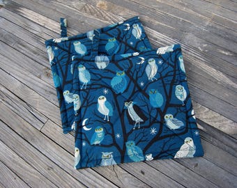 May include: Two square quilted pot holders featuring a dark blue fabric with a repeating pattern of light blue and white owls perched in black tree branches, accented with small white stars and crescent moons, and a fabric hanging loop.