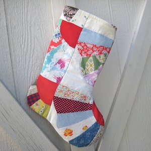 May include: A handmade Christmas stocking with a patchwork design. The stocking features various fabric patterns, including floral, polka dots, and plaid, in red, blue, green, and white. The stocking is hanging against a white wooden background.