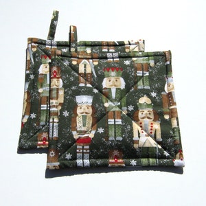 May include: Two green quilted pot holders with a Christmas nutcracker pattern. The nutcrackers are wearing red, blue, and gold hats and have white beards. The background is a green and white snowflake pattern.