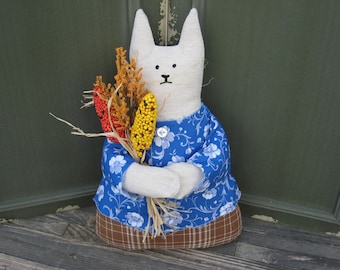 May include: A stuffed fabric cat figure with a white head and body, wearing a blue floral shirt with a single white button and a brown plaid skirt. The cat holds a bundle of dried yellow and red grain stalks in its arms, featuring stitched eyes and nose.