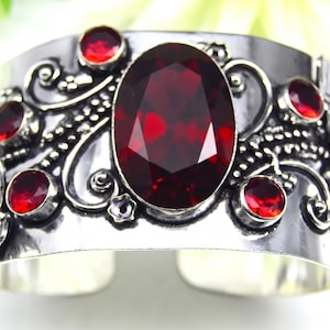 May include: A wide silver cuff bracelet featuring a large central oval-shaped deep red gemstone. The metal surface is decorated with intricate swirling filigree patterns, small floral accents, and several smaller round red stones.