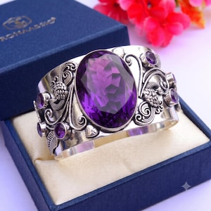 May include: A wide silver cuff bracelet featuring a large, oval-cut, faceted purple amethyst stone at the center. The metal surface is decorated with intricate scrollwork, leaf motifs, and small bird figures, accented by several smaller round amethyst stones.
