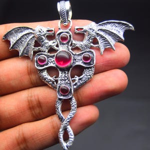 Puede incluir: Un colgante plateado en forma de cruz, con alas y cabezas de dragón en la parte superior. La cruz está adornada con piedras preciosas rojas. El colgante tiene un diseño retorcido en forma de serpiente en la parte inferior.