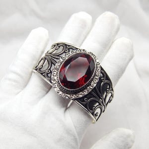 May include: A wide silver cuff bracelet featuring an oval-shaped, faceted deep red gemstone at the center. The metal surface is decorated with intricate, dark-oxidized scrollwork patterns and a beaded border surrounding the central stone.