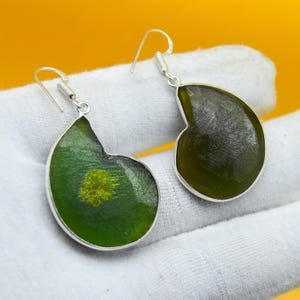May include: A pair of handcrafted earrings with a unique design. Each earring features a dark green, teardrop-shaped pendant with a silver-colored border. The pendant has a lighter green and yellow center, and a silver hook.