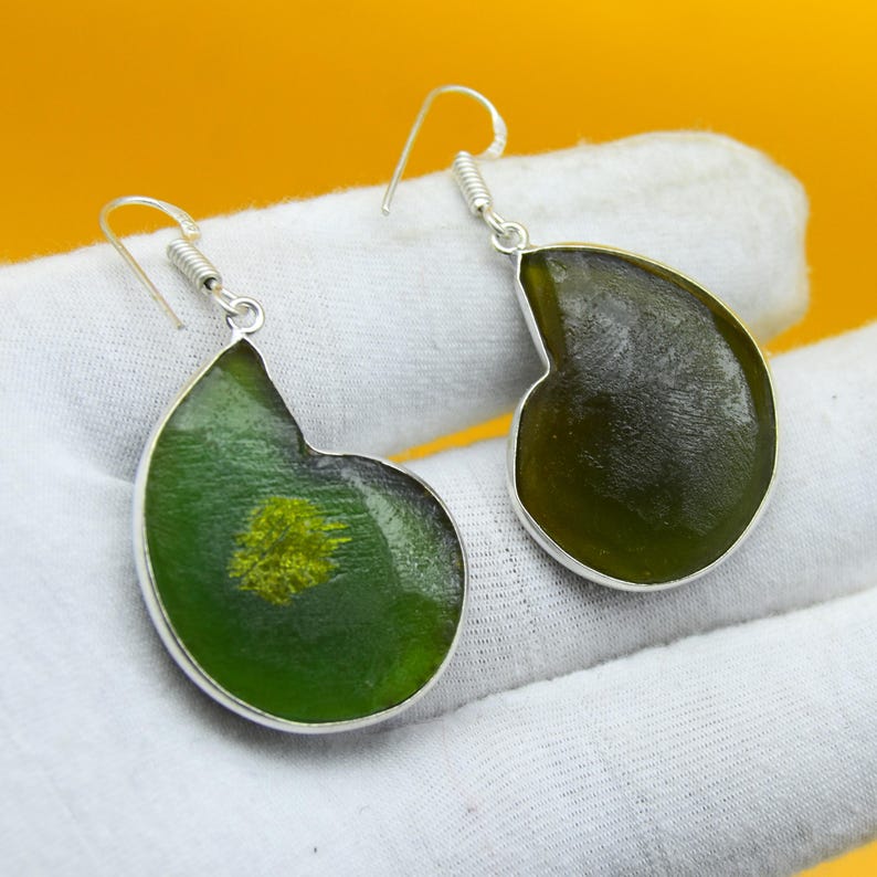 May include: A pair of handcrafted earrings with a unique design. Each earring features a dark green, teardrop-shaped pendant with a silver-colored border. The pendant has a lighter green and yellow center, and a silver hook.