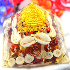 May include: A clear resin pyramid with a gold-colored structure at the top. The pyramid is filled with seashells, red and brown organic shapes, and a copper spiral. The background features blurred red and blue flowers.