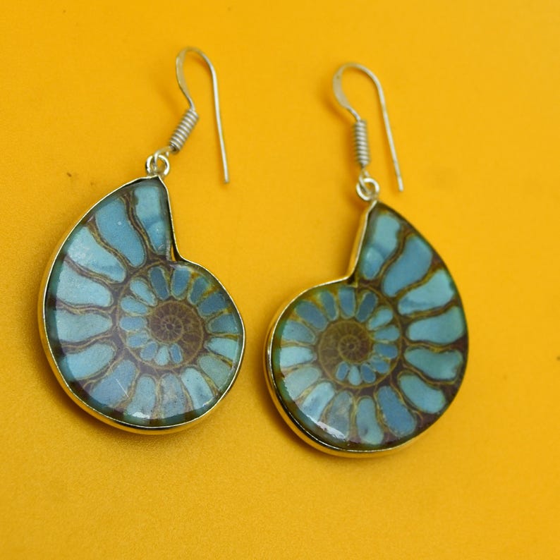 May include: A pair of silver-toned earrings featuring a fossilized ammonite design. The earrings have a blue and brown color scheme, with a spiral pattern. The earrings are set against a yellow background.