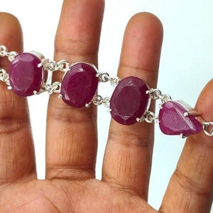 Puede incluir: Una pulsera de plata con grandes piedras preciosas rojas oscuras de forma ovalada. Las piedras están conectadas por eslabones de plata y tienen un cierre de palanca. La pulsera se muestra sobre un fondo claro.