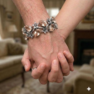 May include: A silver bracelet with large, oval, peach-colored gemstones and small flower accents. The bracelet is worn on a wrist, with another person's hand holding it. A silver ring is visible on one finger.