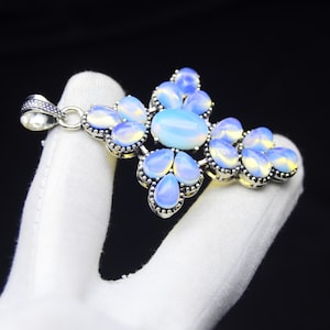 Puede incluir: Colgante plateado con forma de cruz, engastado con piedras opalescentes. El colgante tiene una piedra central ovalada y numerosas piedras en forma de lágrima en tonos azules y blancos. El colgante está sujeto por una mano enguantada de blanco sobre un fondo negro.